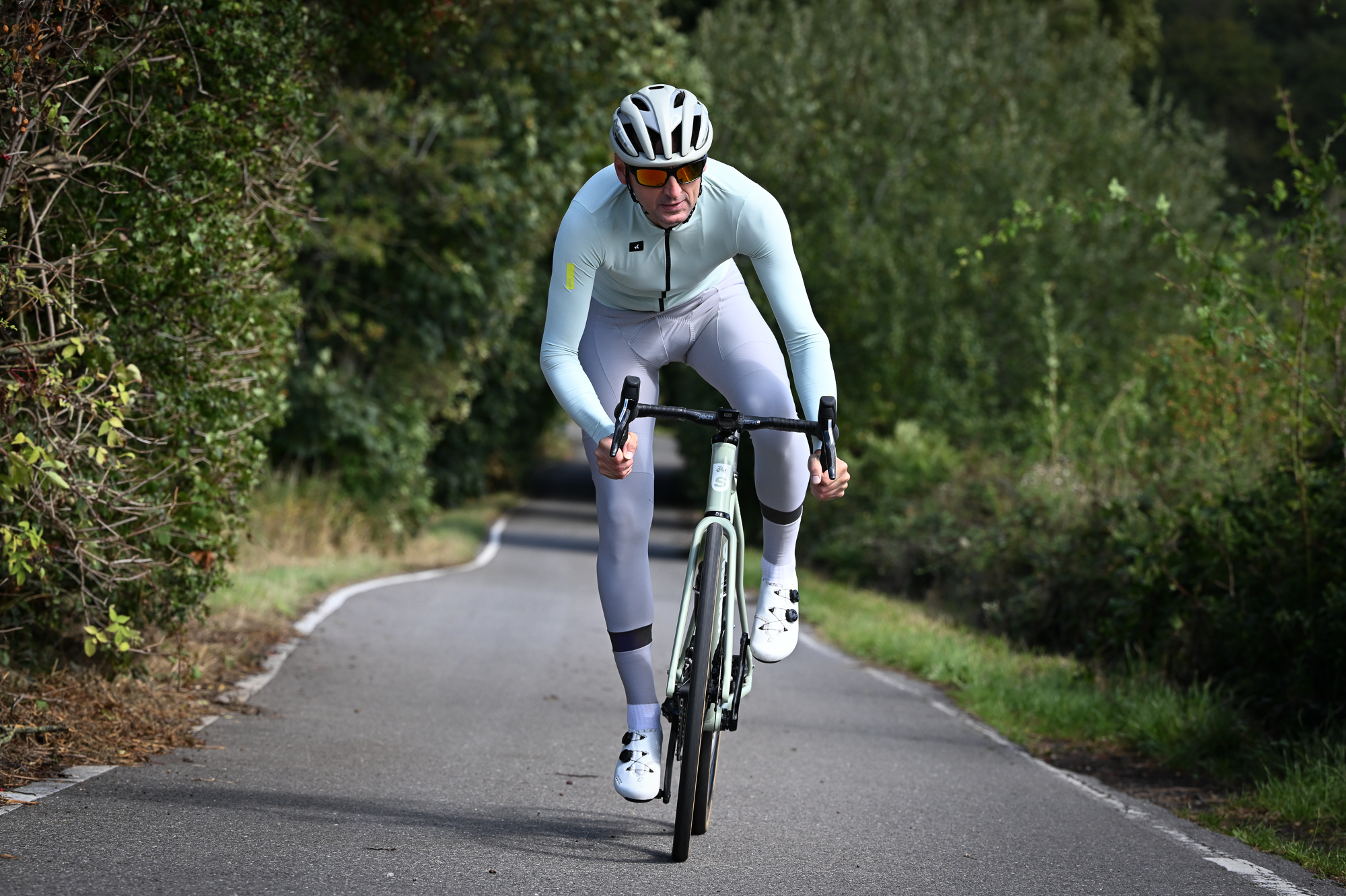 Man riding out of the saddle towards the camera, wearing a pale green long sleeved jersey and pale grey bib tights