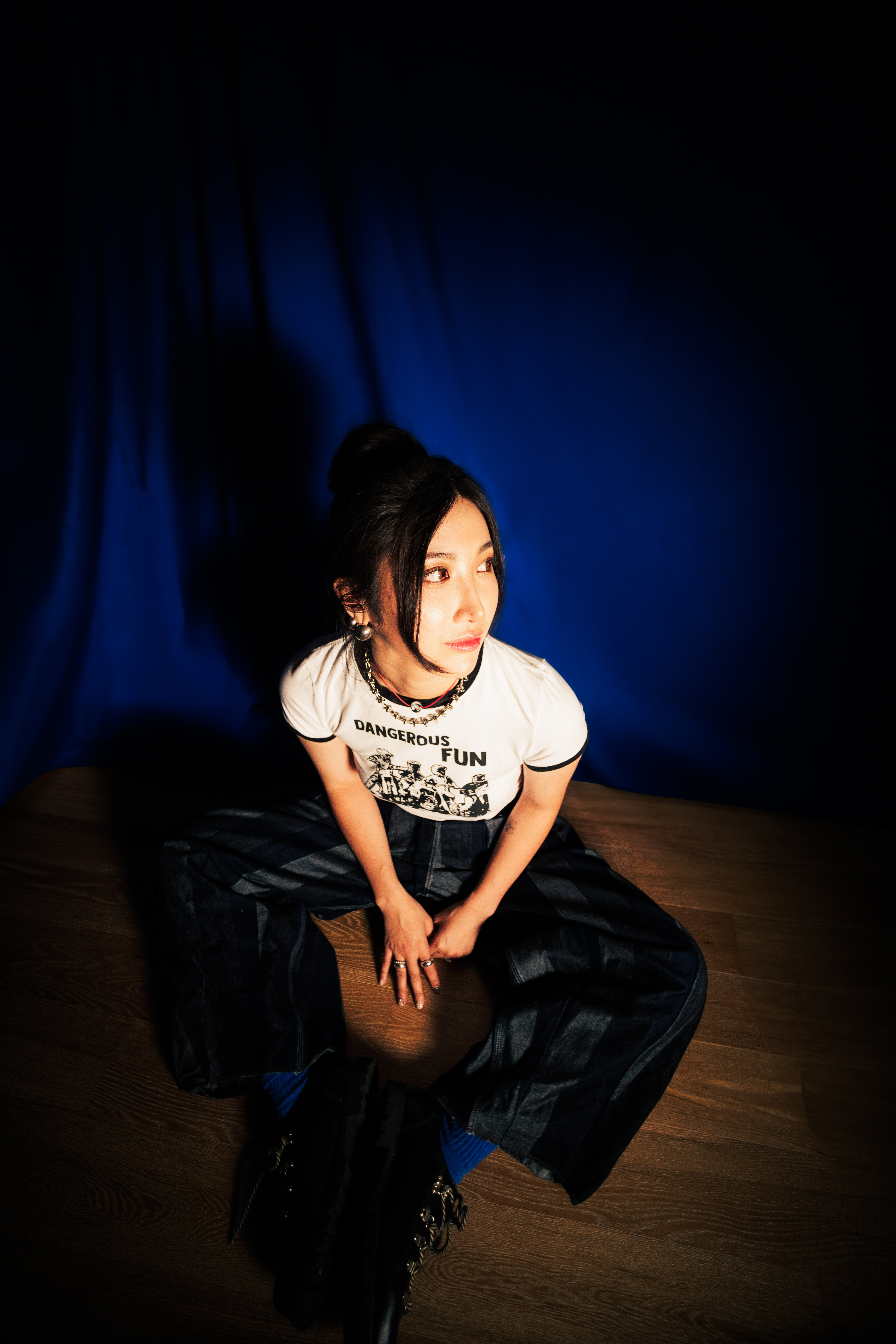 A portrait of singer-songwriter EJAE, wearing a white t-shirt and black pants while sitting on a wooden floor in front of a blue backdrop.