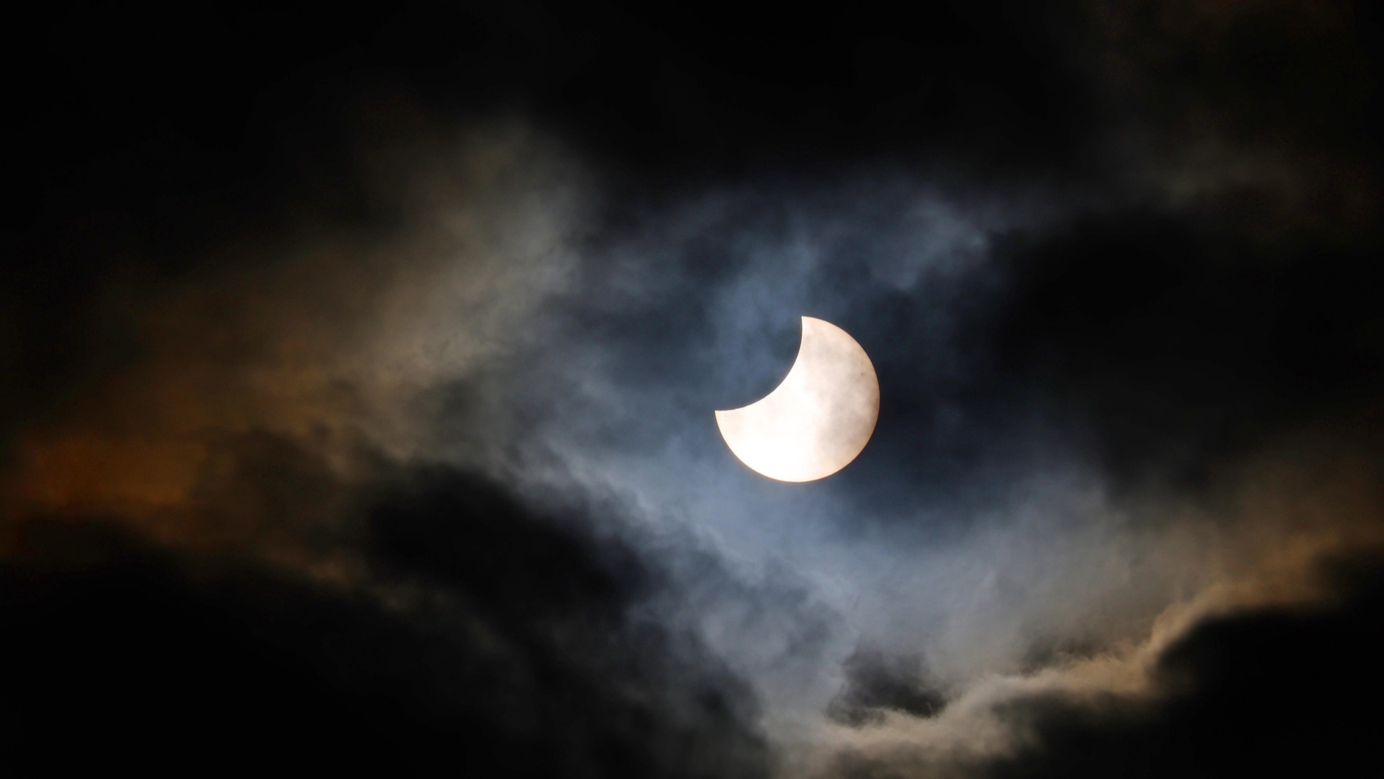 partial solar eclipse appearing from behind some broken cloud.