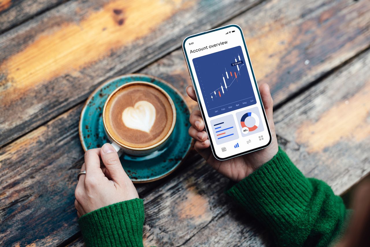 Overhead view of young woman checking stock market data on smartphone while drinking coffee