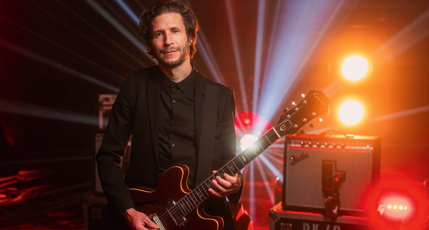 A portrait of Interpol guitarist Daniel Kessler pictured with his rig. He plays an Epiphone Casino and there is a Fender Princeton Reverb in a background lit with orange, red and white stagelights.