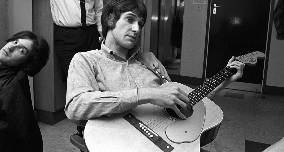 A black-and-white image of Ray Davies of the Kinks plays a Fender Malibu backstage with his bandmates in the background