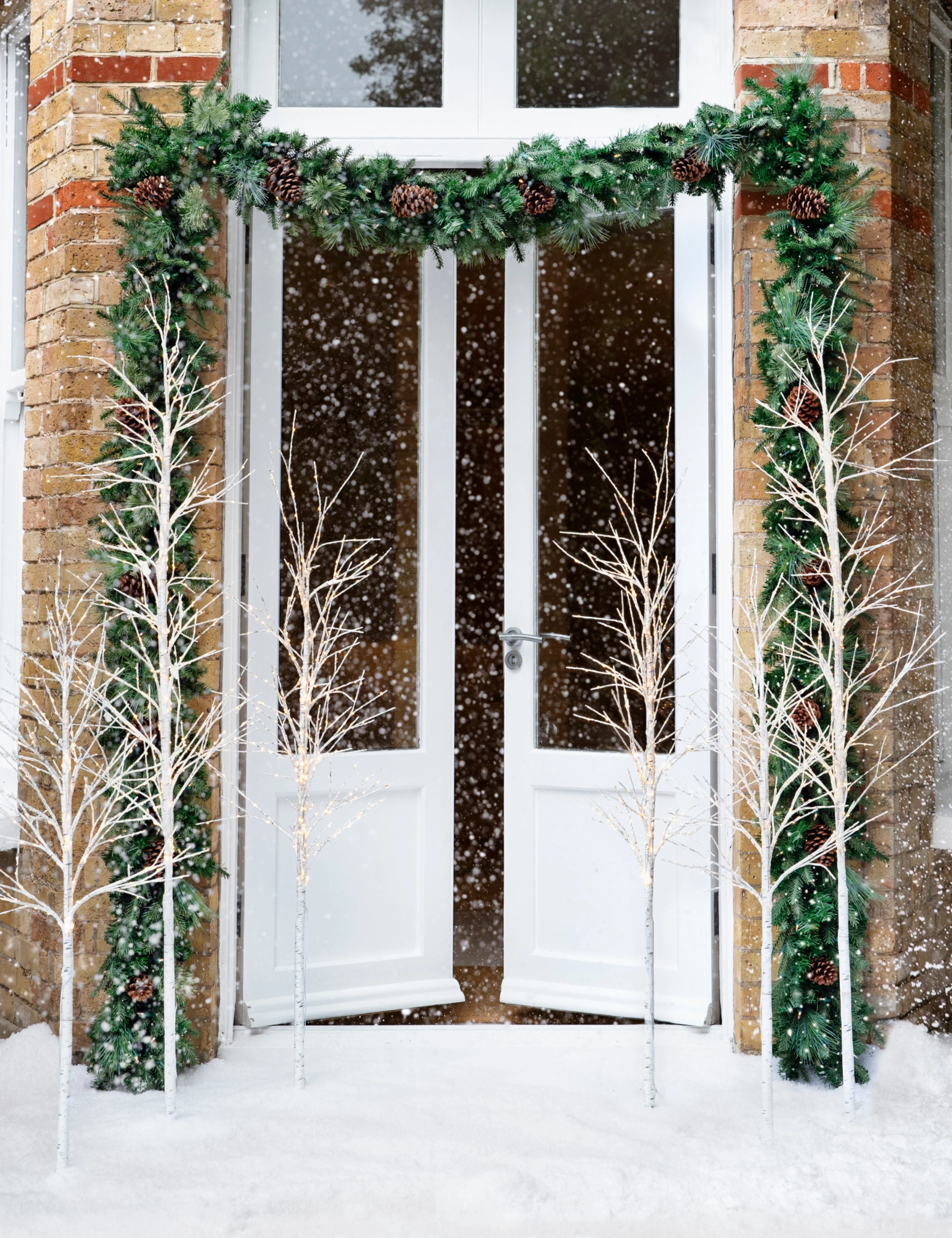 Twinkling twig trees flanking either side of a grand brick entryway, which is draped with a swag of evergreen garland.