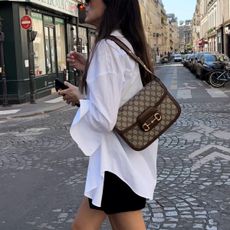 Paris-based model and content creator Leia Sfez wearing a white poplin button-down shirt with a Gucci Horsebit bag and black shorts walking in the summer.