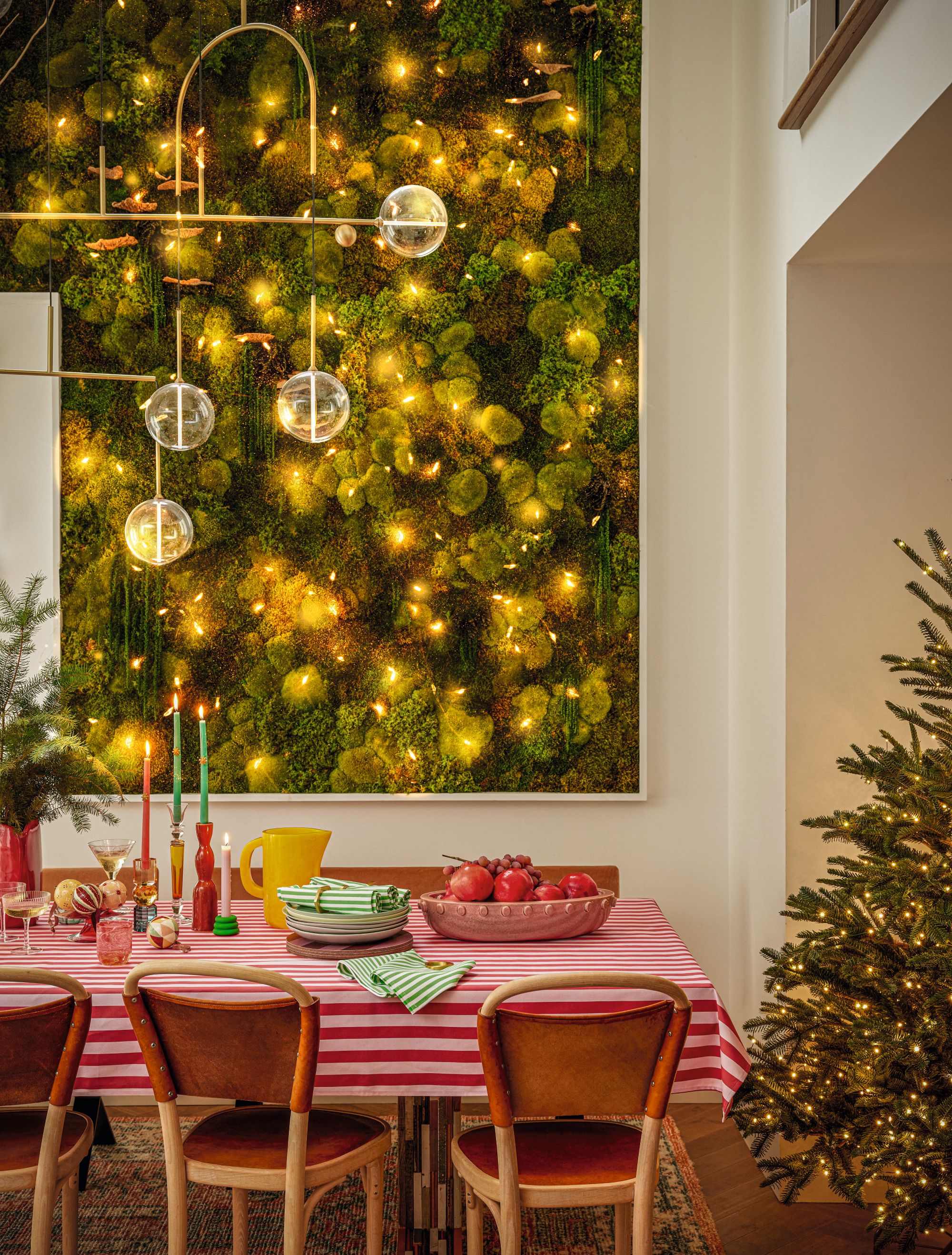 A Christmas tree is half in view next to a table with a red and white striped tablecloth with some fruit, candles, napkins, a jug and a vase with christmas tree branches in; there's also an abstract gold and white globe pendant light hanging from the ceiling and a lit artificial or real living wall in the background