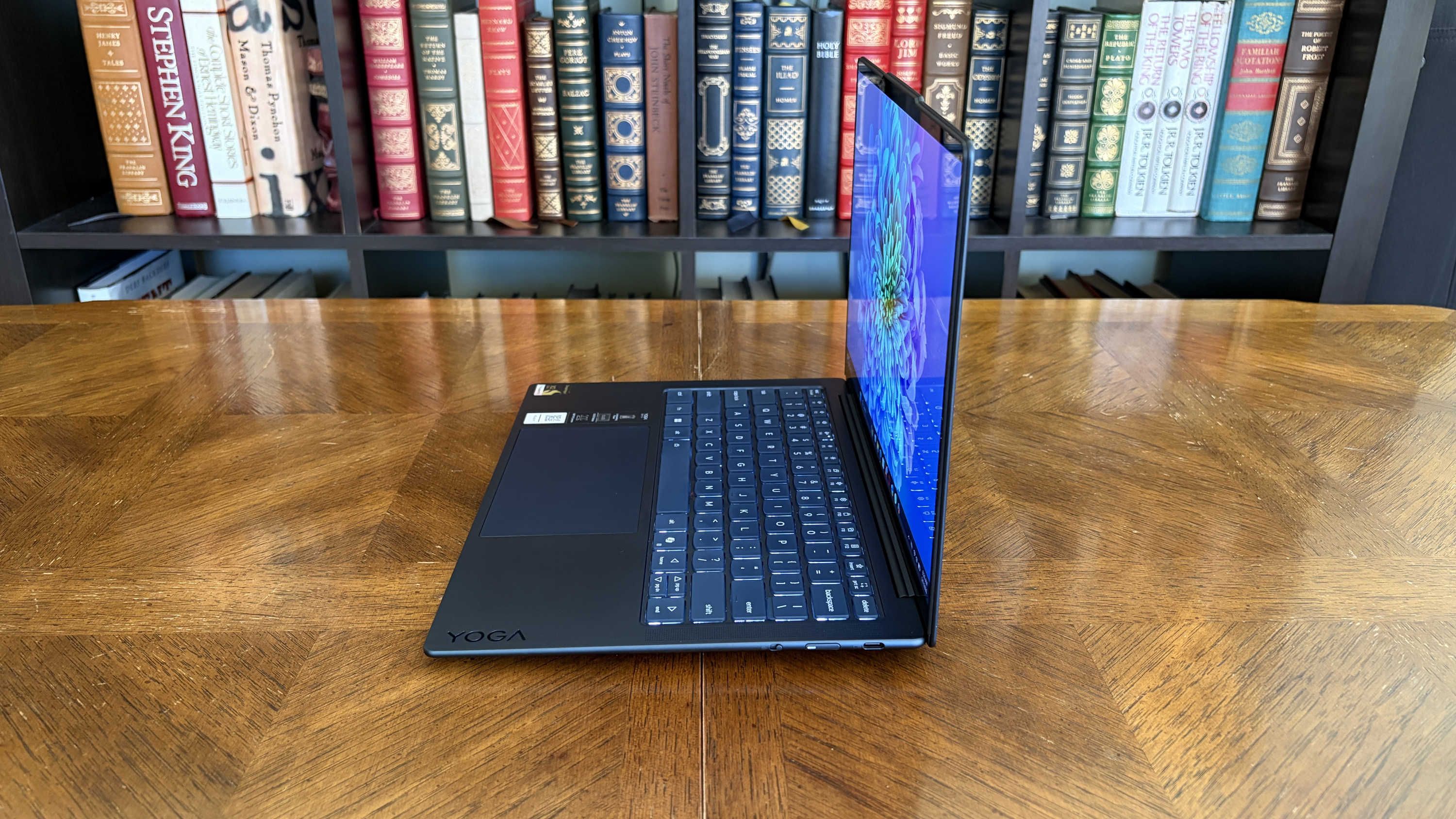 A sleek laptop, partially open, sits on a wooden table. The screen displays vibrant colors. Behind are shelves filled with assorted books, creating a scholarly atmosphere.