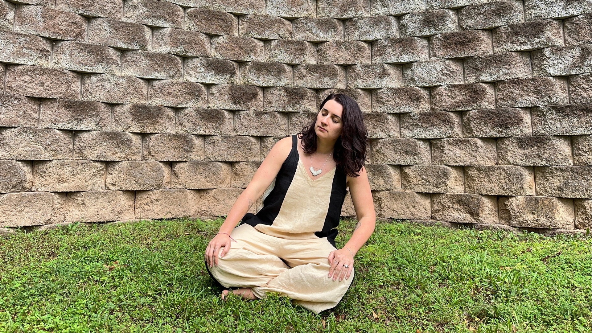 woman sitting cross legged rotating around her hips. she's on some grass, outside, in front of a large stone wall