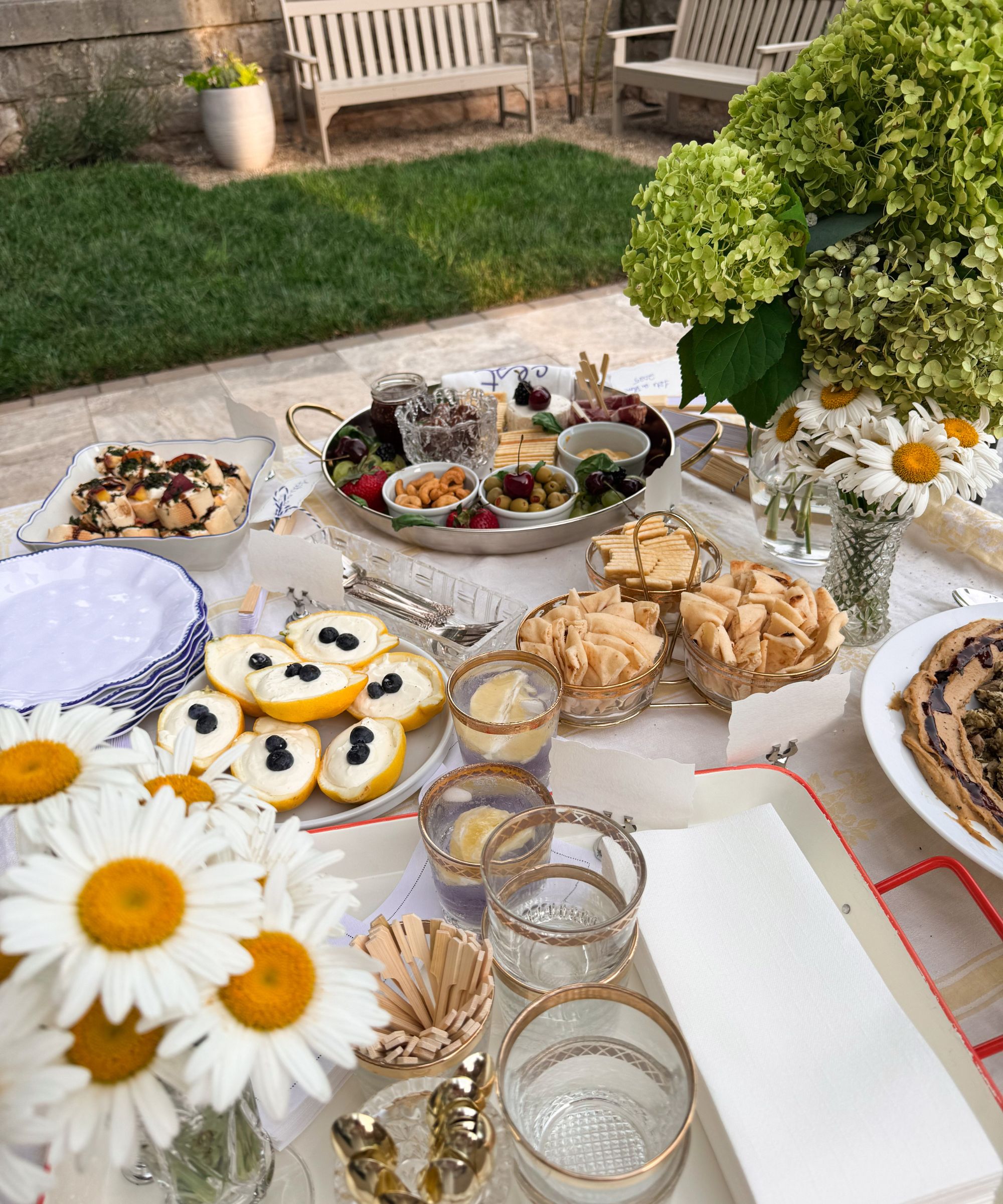 Summer dining table with thrifted silver platters, vintage glassware, bowls of snacks and desserts, and a vase of daisies and hydrangeas