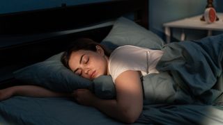 A woman with dark hair sleeps in bed on a dark winter morning