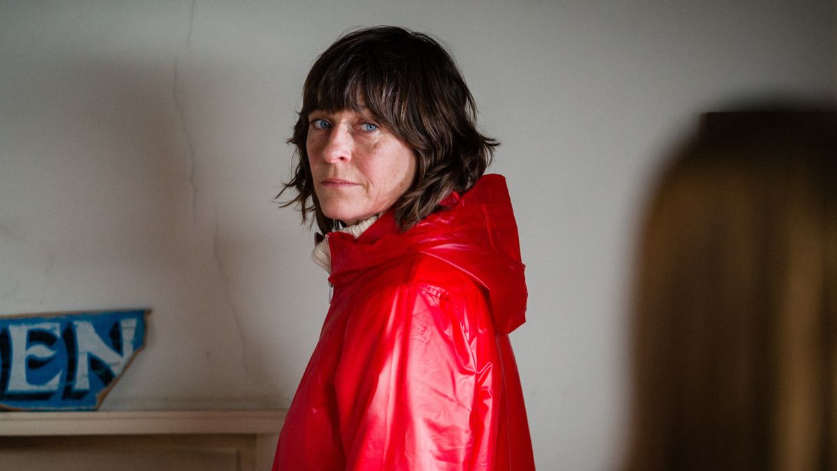 A woman in a red mac standing in front of a mantelpiece. 