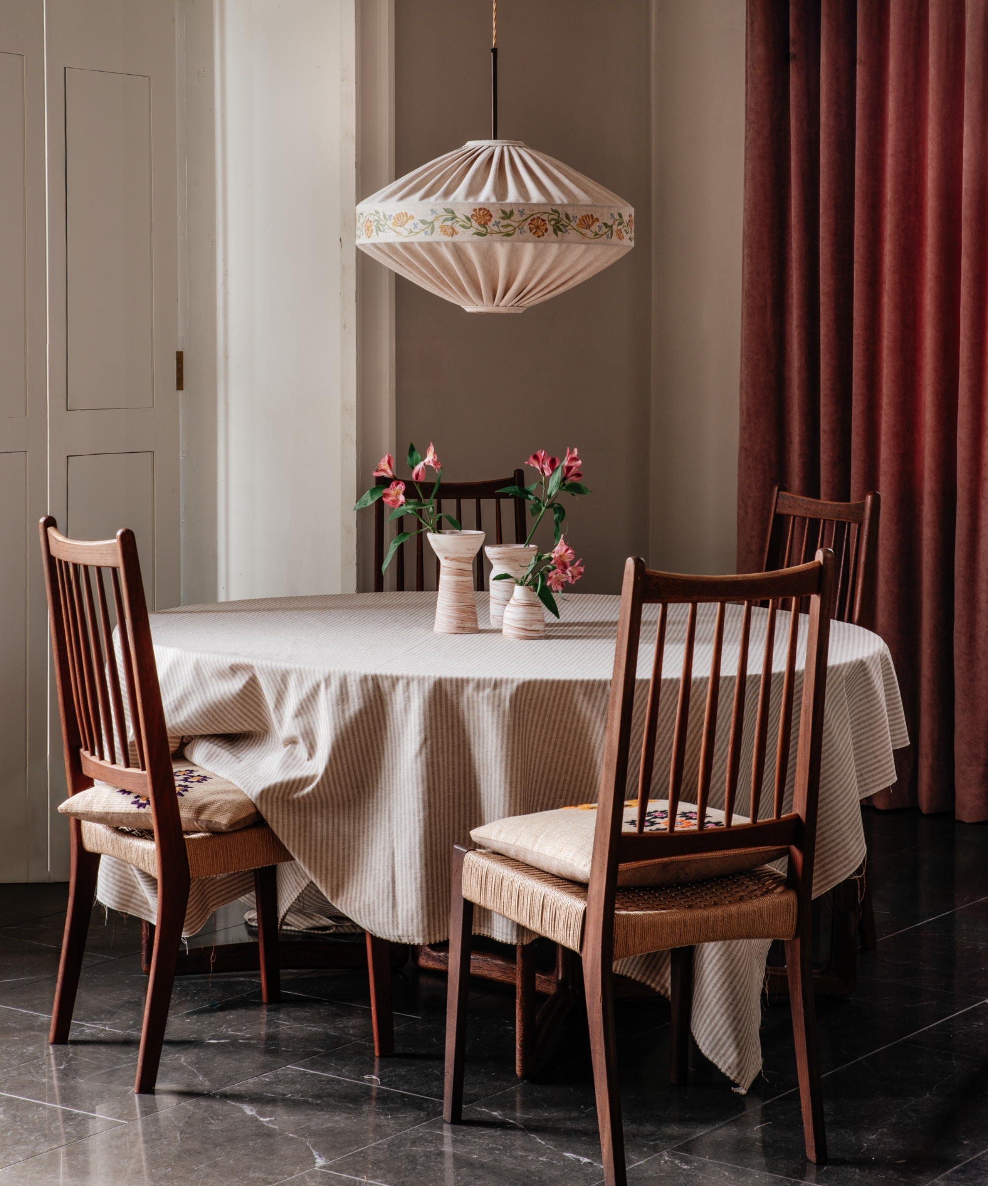 A sculptural pleated linen pendant light with floral embroidery hangs above a dining table with wooden chairs and pink flowers.