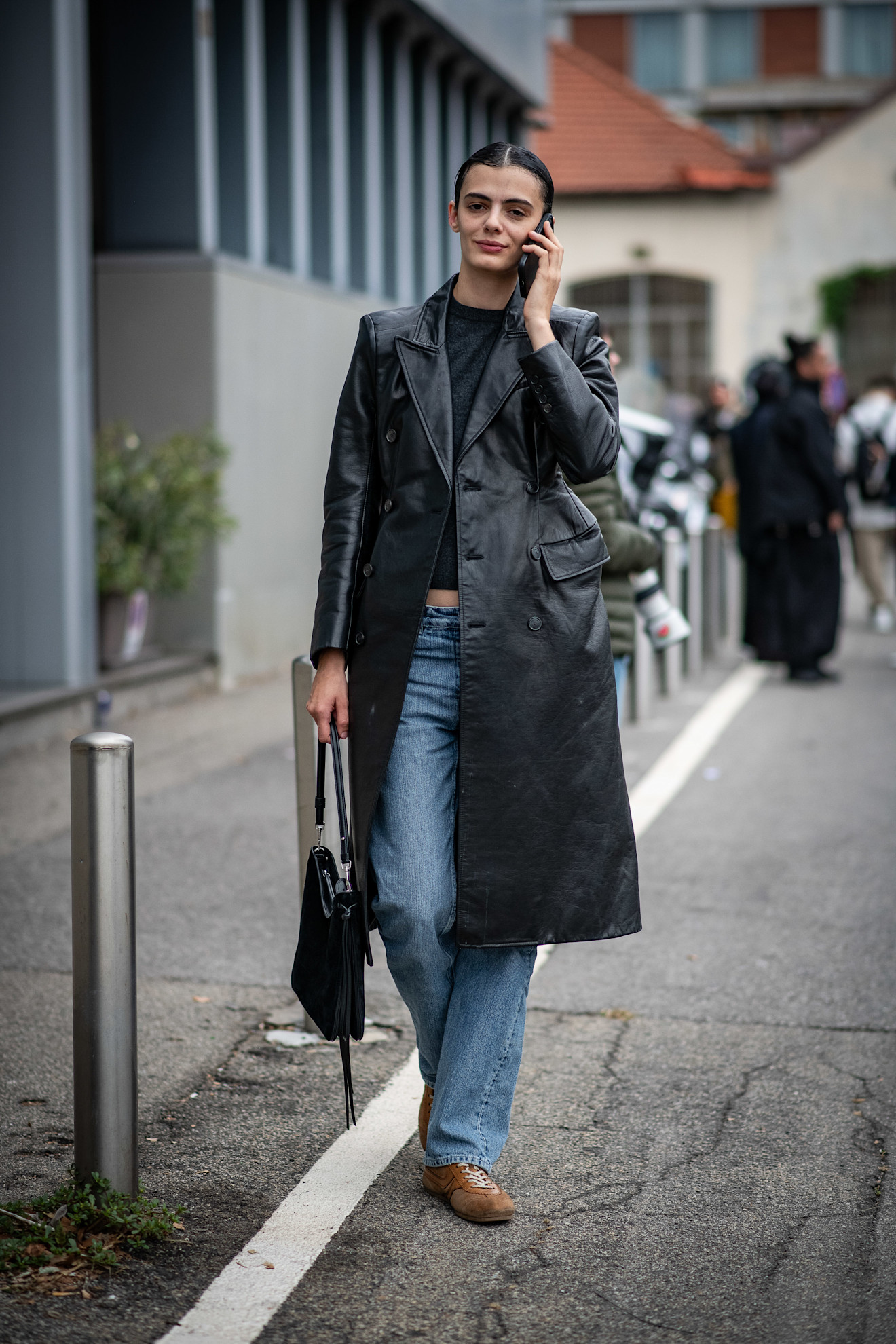 fashion week attendee talking on the phone wearing leather trench coat, jeans, and brown sneakers