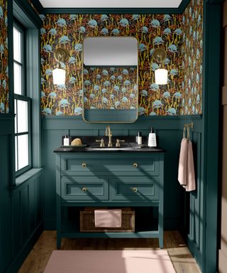 A bathroom with forest green panels and bathroom vanity, wallpaper with a blue and gold floral patterned wallpaper, and a gold rectangular mirror and towel hook