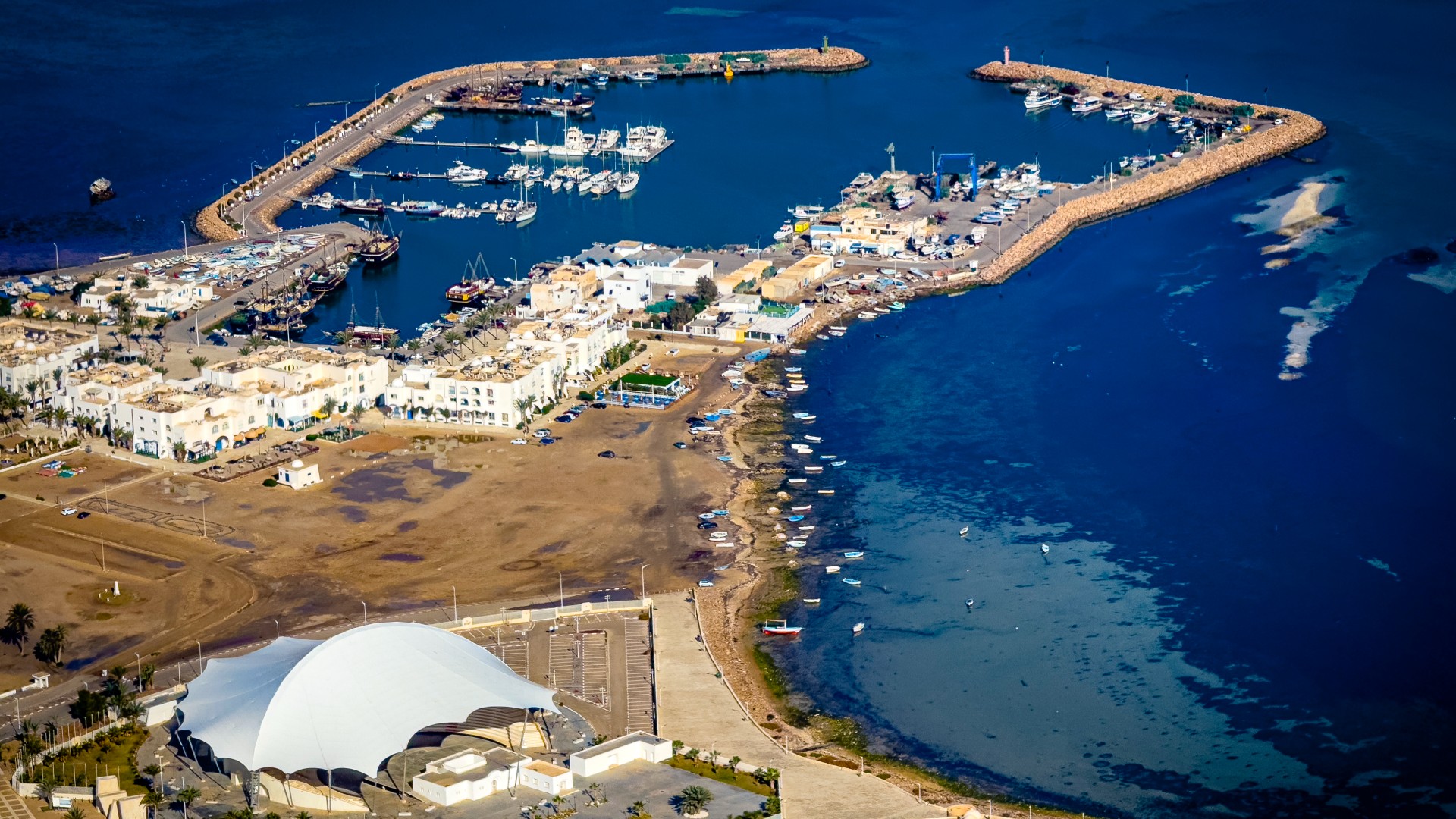 Aerial view of a coastal town featuring a marina with docked boats, a large white amphitheater, and an ancient stone fortress near the water.