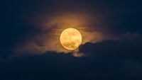 An orange full moon is pictured glowing in a cloudy dark night sky.