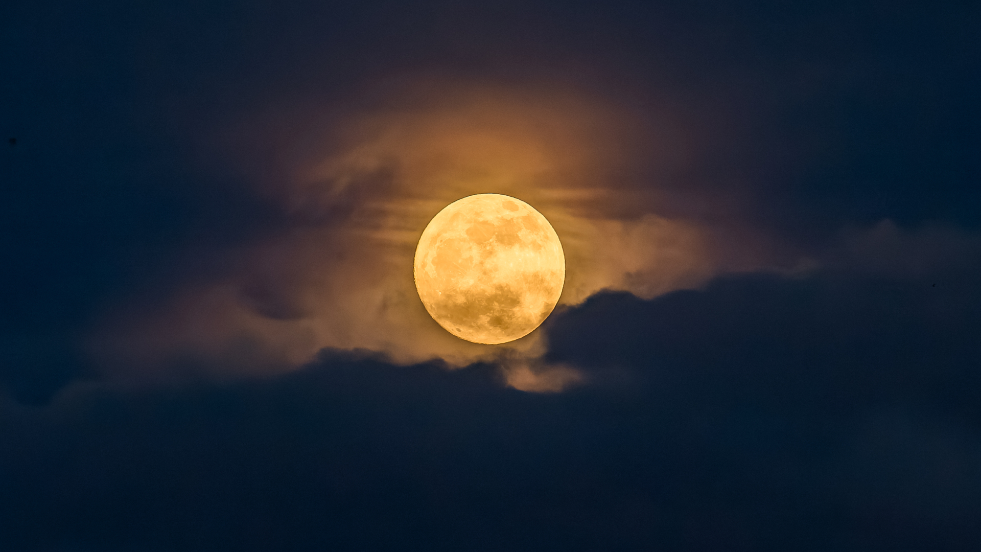 An orange full moon is pictured glowing in a cloudy dark night sky.