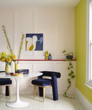 Dining room with bright yellow feature wall, other wall with wooden paneling painted white, red painted built-in wooden shelf, white knoll saarinen oval dining table, upholstered blue velvet dining chair, abstract blue, white and yellow artwork and decorative accessories