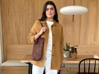 British fashion influencer Anna Howard poses in her modern wood-paneled London kitchen wearing a tan suede jacket, burned-out sheer t-shirt, brown woven tote, and white jeans