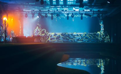 Stage for the Absolut Art Awards, blue and orange spotlights shone on the artwork, ceiling lights on metal frame, dark steps surrounding a pool of water reflecting the stage