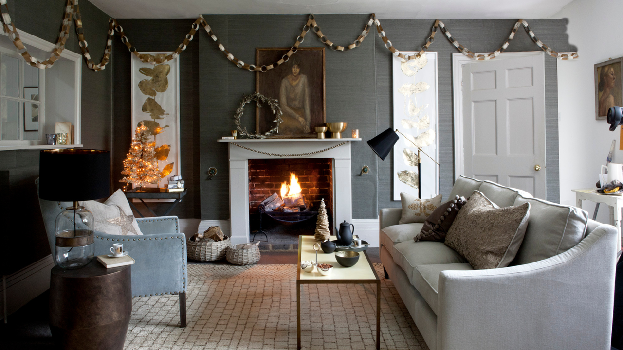 Grey living room, cream three seater sofa, silver and cream cushions, blue studded armchair, drum stool table, coffee table, glass jar base lamp, open lit fireplace, wicker log baskets, gold leaf print framed artwork, paper chains, small paper Christmas tree