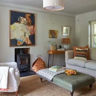 sitting room with pale sage green walls and large artwork above woodburning stove, green upholstered coffee table footstool