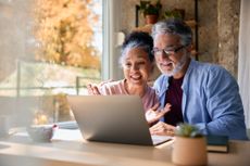 Cheerful mature couple waving at the laptop making a video call with their family while sitting at the kitchen table, enjoying a healthy breakfast.
