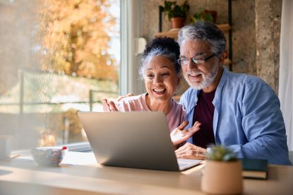 Cheerful mature couple waving at the laptop making a video call with their family while sitting at the kitchen table, enjoying a healthy breakfast.