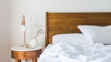 Should you turn your AC off at night? Here is a cool white bedroom with a bed with a dark wooden headboard with white pillows and sheets on the mattress and a white side table with a wooden base, a rose gold lamp, and a white vase with green stems in it