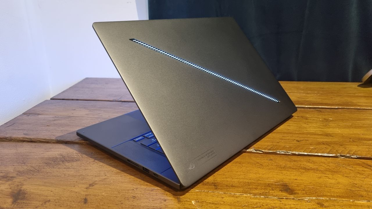 The Asus ROG Zephyrus G16 gaming laptop open on a wooden desk shot from behind, showing the rear lighting