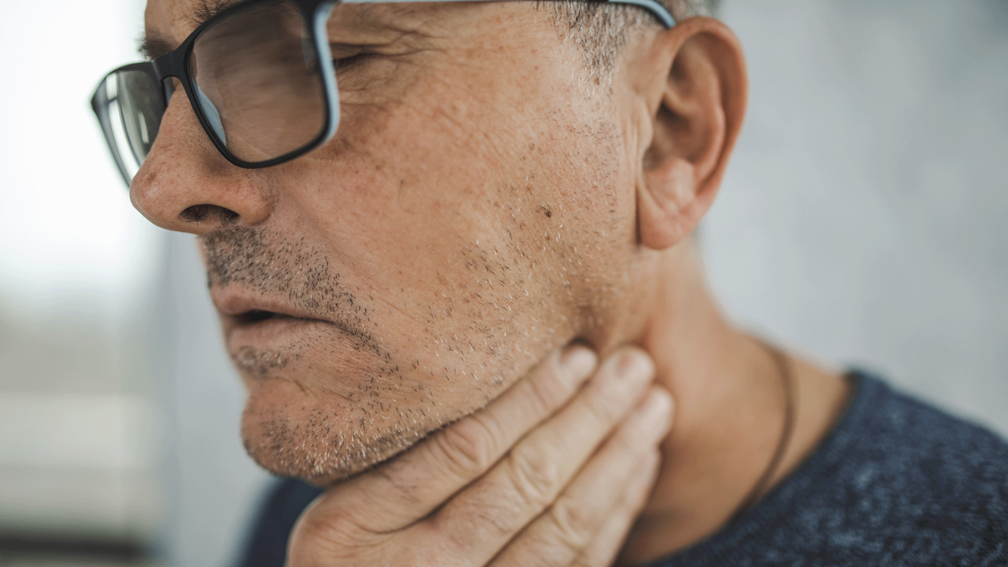 An older man with gray stubble and glasses clutches his throat
