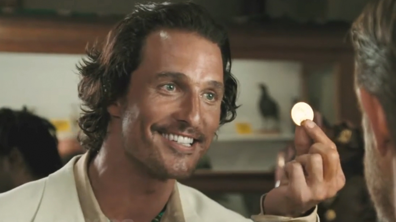 Matthew McConaughey smiling and holding up a gold coin in Sahara