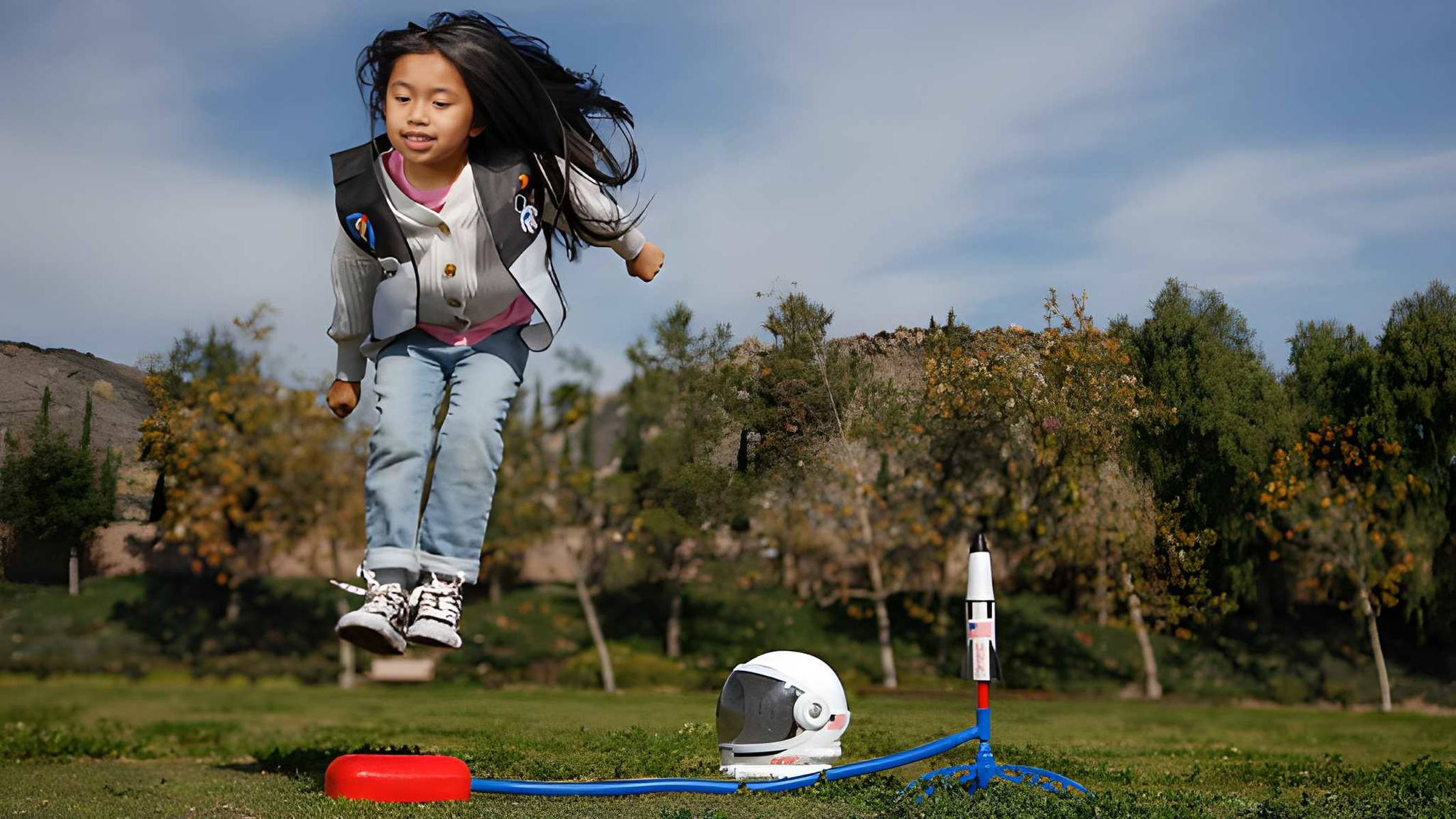 A girl leaping onto a stomp rocket's launcher, with a toy astronaut helmet nearby.