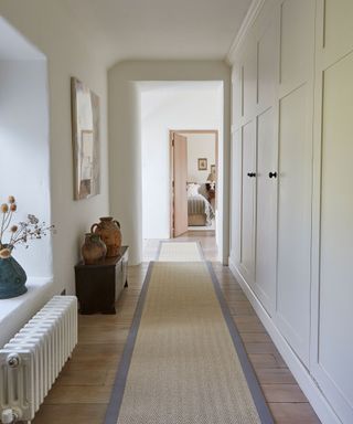 Country style hallway with white painted walls, built in floor to ceiling cupboards, wooden floorboards with natural and light gray runner, low radiator beside window with blue vase and dried flowers on windowsill, dark wooden chest with traditional vases, looking out onto bedroom with wooden door and doorframe