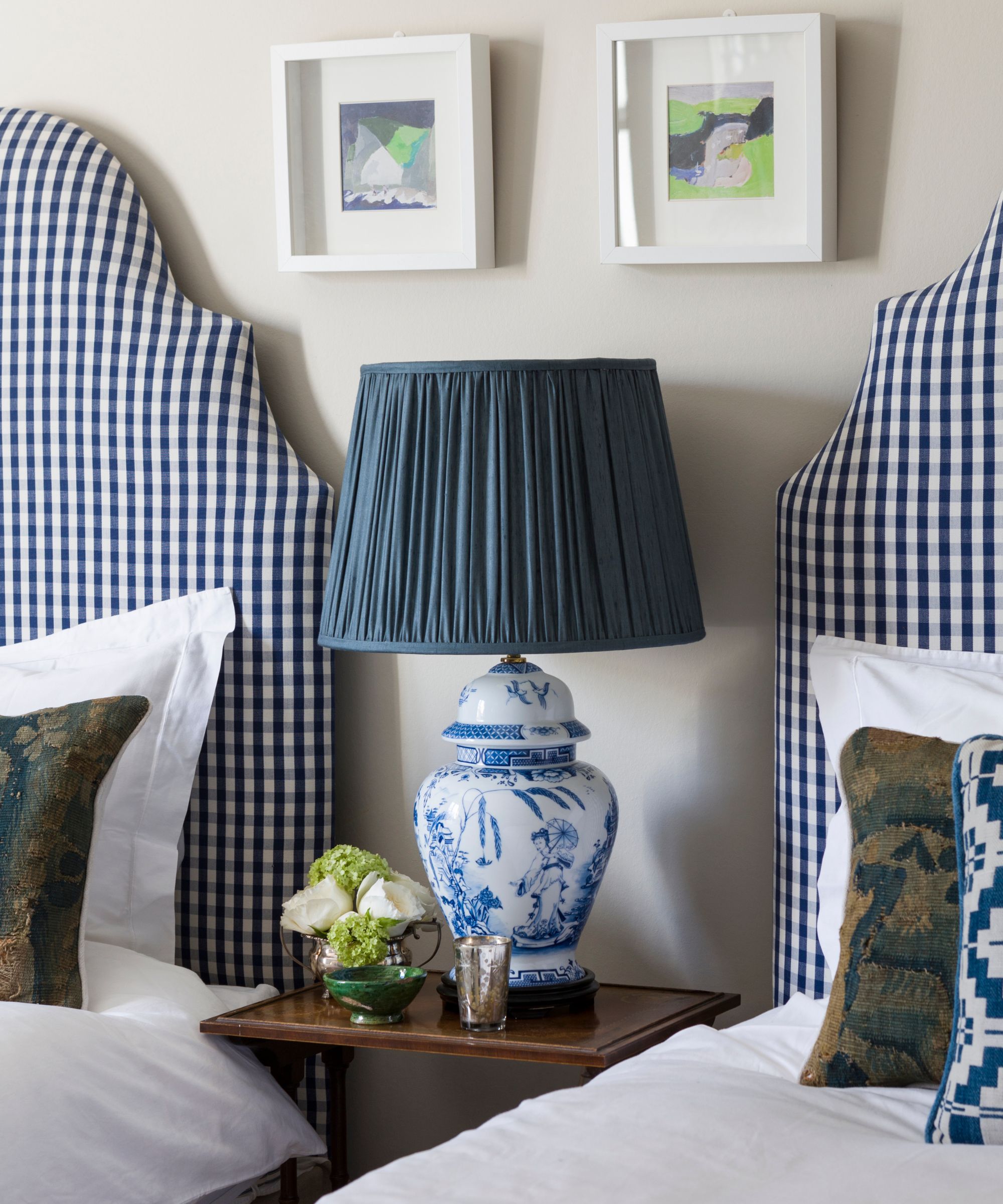 A white bedroom with matching twin beds with gingham headboards, with a brown wooden bedside table between. On top of the bedside table is a ceramic decorative lamp with a teal lampshade, and above are two framed artworks.