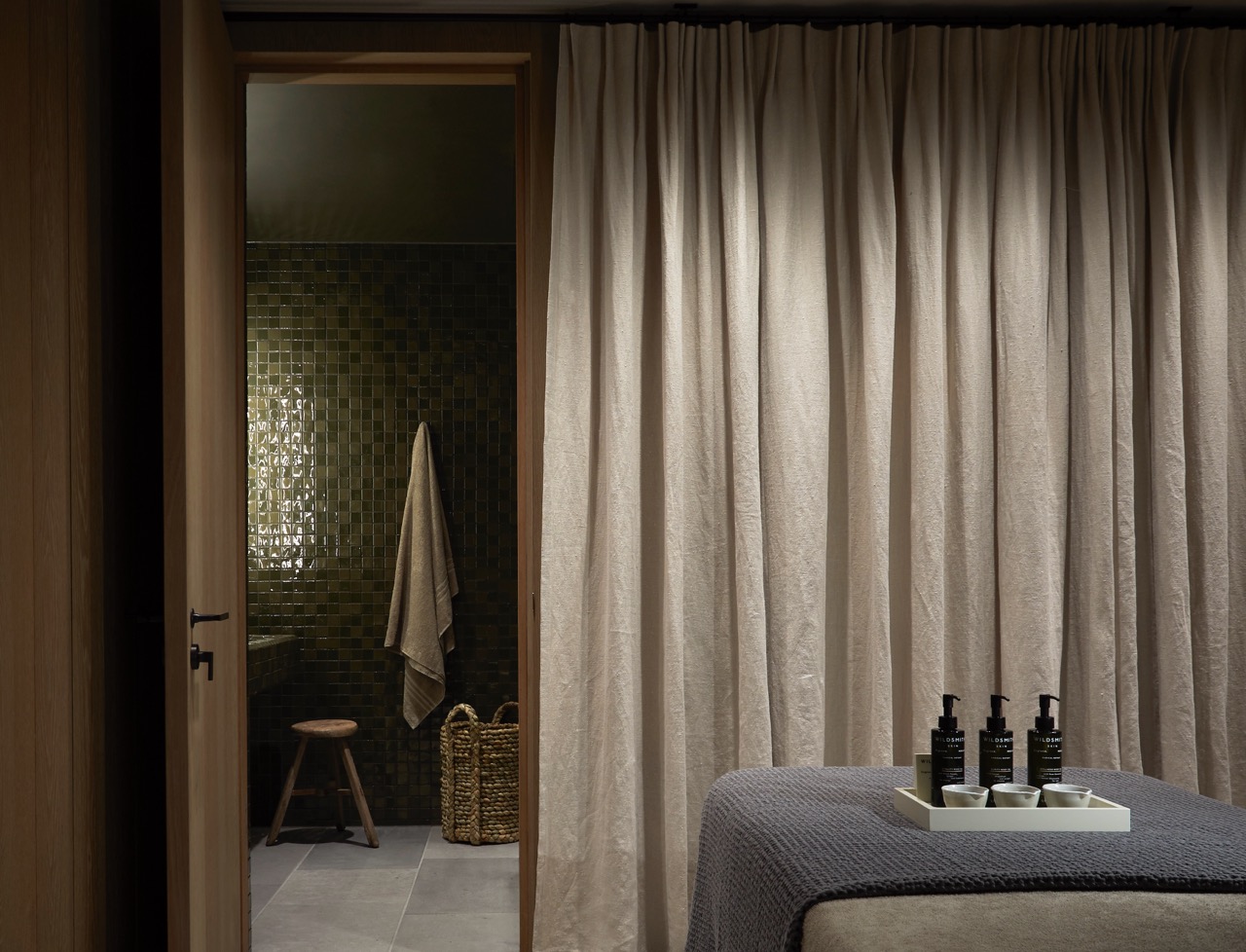 A dimly lit treatment room with a massage bed draped in grey fabric, bottles of Wildsmith products, and a glimpse of a tiled green bathroom through an open door.