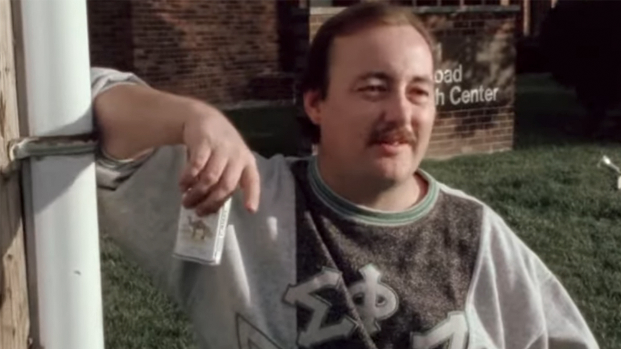 A man holding a pack of Camel cigarettes and leaning against a pole in Roger & Me