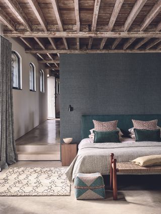 contemporary bedroom with grey textured wallpaper, beamed ceiling, green headboard, rug, stone floor
