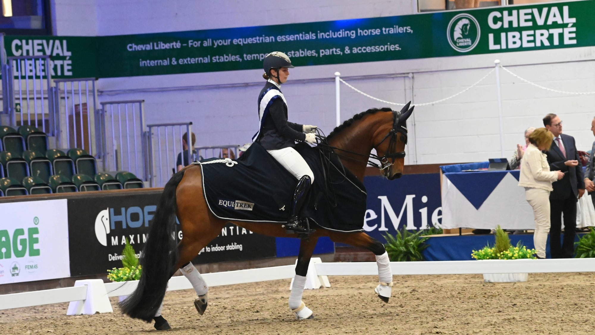 Woman rides a bay horse in an indoor arena. The rider wears a white winner&amp;rsquo;s sash and the horse wears a blue winner&amp;rsquo;s rug at the winter dressage championships.