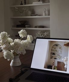 Image of laptop on dark wood table with vase of flowers next to it, laptop shows an image of a shelf that can also be seen in the corner of the room