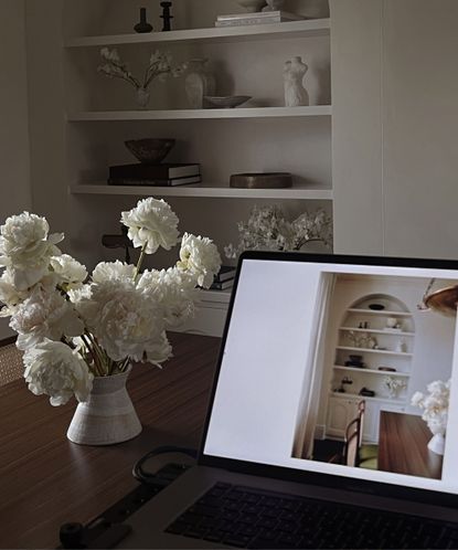Image of laptop on dark wood table with vase of flowers next to it, laptop shows an image of a shelf that can also be seen in the corner of the room