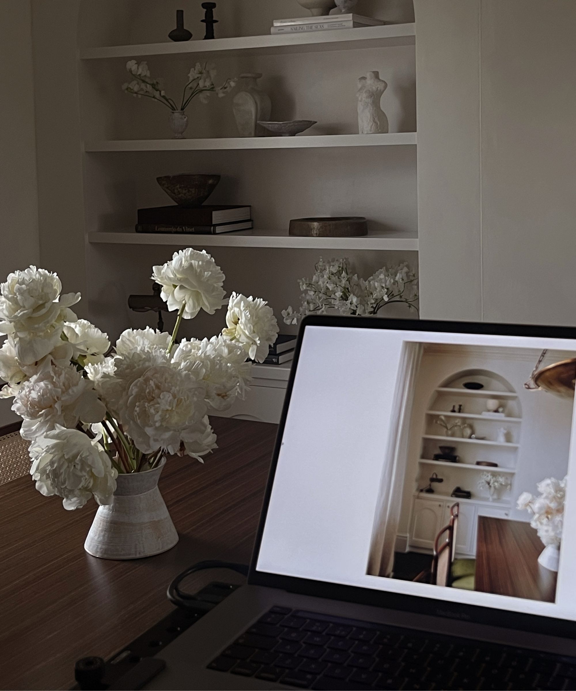 Image of laptop on dark wood table with vase of flowers next to it, laptop shows an image of a shelf that can also be seen in the corner of the room
