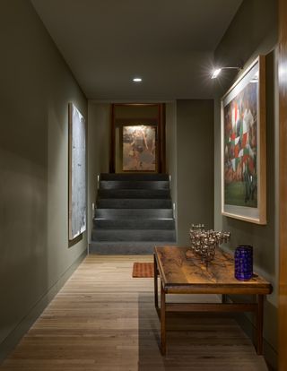 Styled interior at Ventana House in Mexico, Olive walls, wooden floor, natural wood table, wall art , wall light, grey steps, floor mat, wooden door frame, white ceiling, spotlight