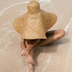 Woman in a straw hat sat on the beach with the sea around her, shot from above