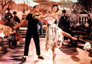 a man in an all black outfit with yellow accents dances with a woman on point in a floral dress in An American in Paris