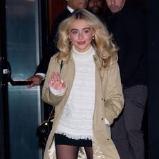 a paparazzi photo of a white woman with blonde hair wearing a beige trench coat and a white turtleneck sweater
