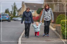 Mother and grandmother walking along the pavement holding hands with younger toddler, who is walking between them