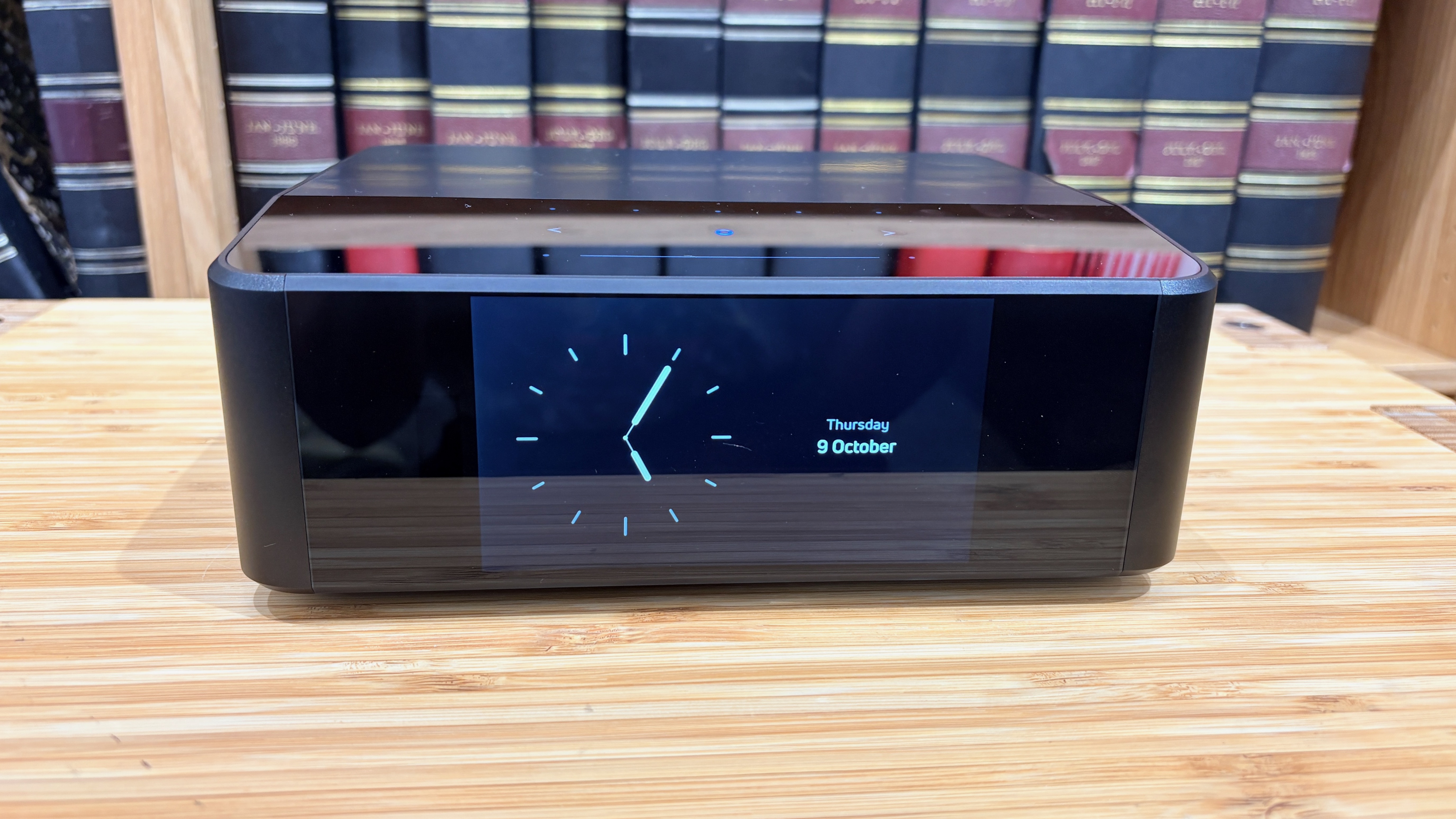 Bluesound Node Icon music streamer on wooden hi-fi rack in front of bookcase, on screen is clock face and date