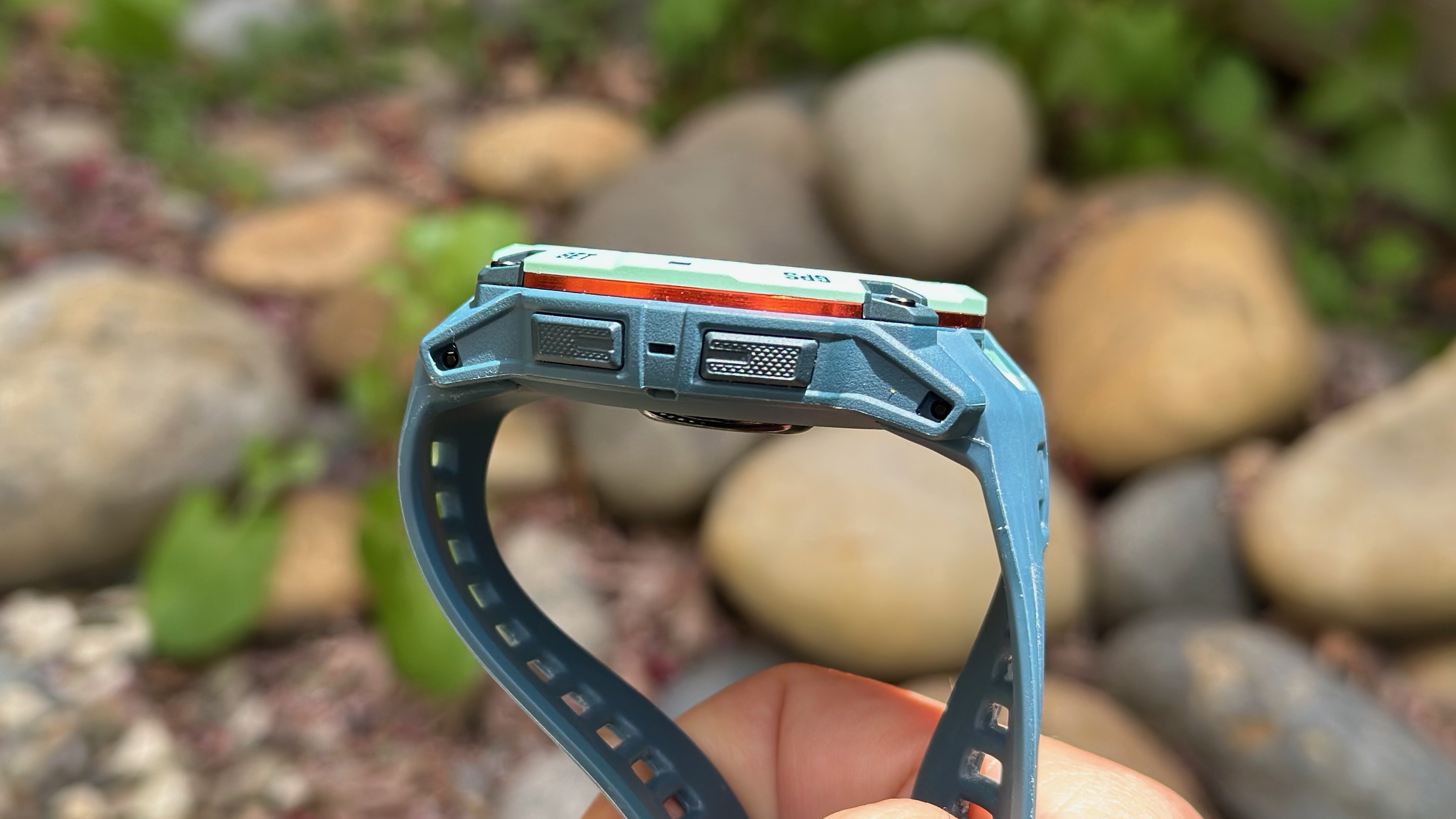The right side of the Garmin Instinct 3 50mm AMOLED, the photo focused on the two buttons and orange aluminum bezel, with blurred rocks in the background.