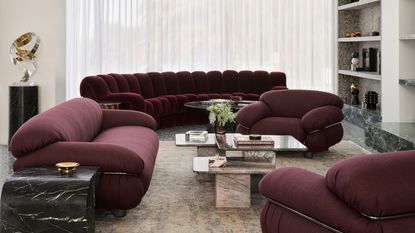 modern living room with arched window near ceiling, sheer curtains, a built-in bookcase wall, patterned rug, stone nesting coffee tables, and a curved velvet purple sofa with purple armchairs and stone side table