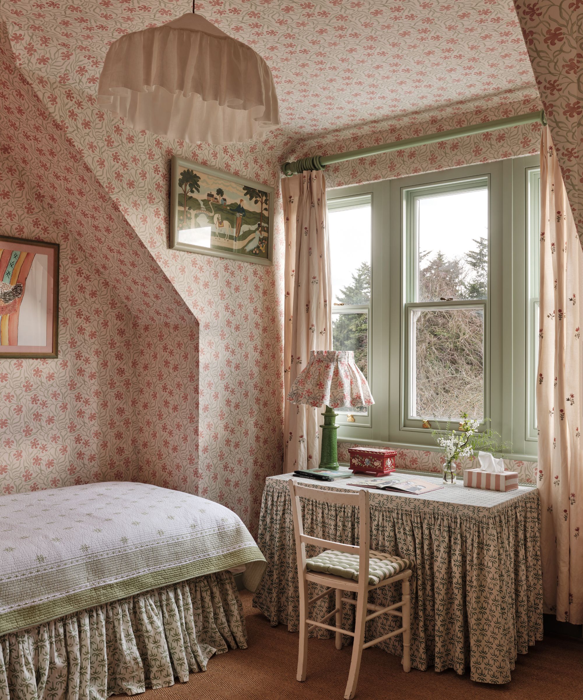 Bedroom with floral wallpapered walls, a skirted bed with a green and white quilt, a desk with skirted fabric draped on top, a sage green painted window, and a pleated pendant light on the ceiling.